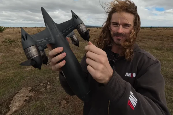 Man holding a black high-speed Blackbird drone outdoors in a grassy field under a cloudy sky, showing the aircraft’s sleek body, twin rear fins, and large propulsion units.