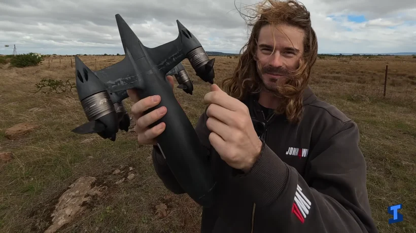 Man holding a black high-speed Blackbird drone outdoors in a grassy field under a cloudy sky, showing the aircraft’s sleek body, twin rear fins, and large propulsion units.