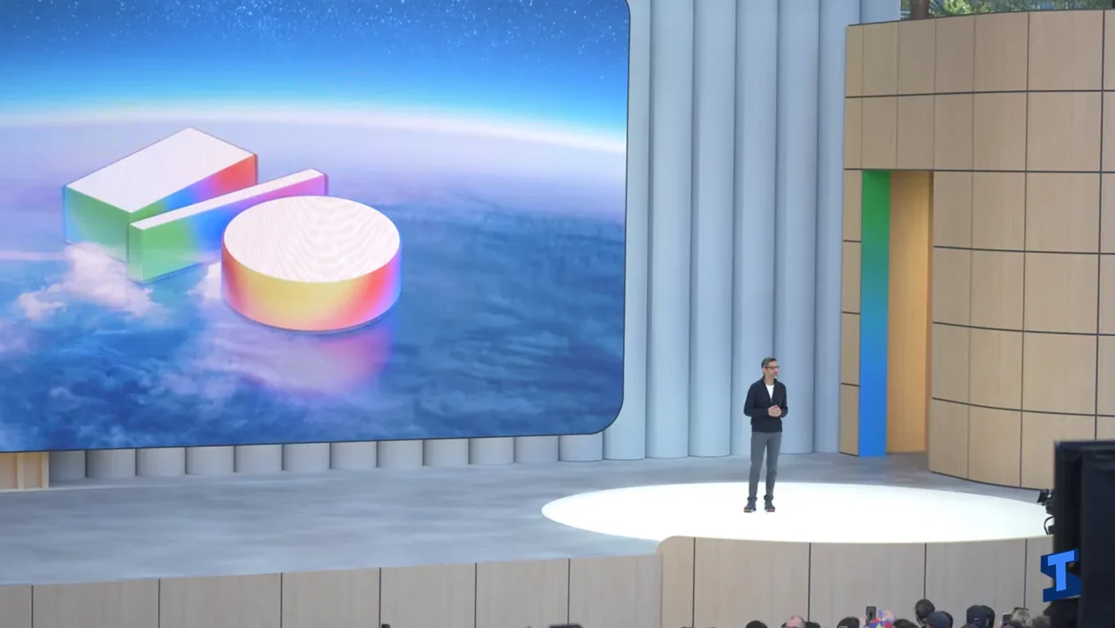 Google I/O keynote stage with a presenter standing under a spotlight beside a large screen showing colorful AI-themed 3D shapes above a cloud-like background.